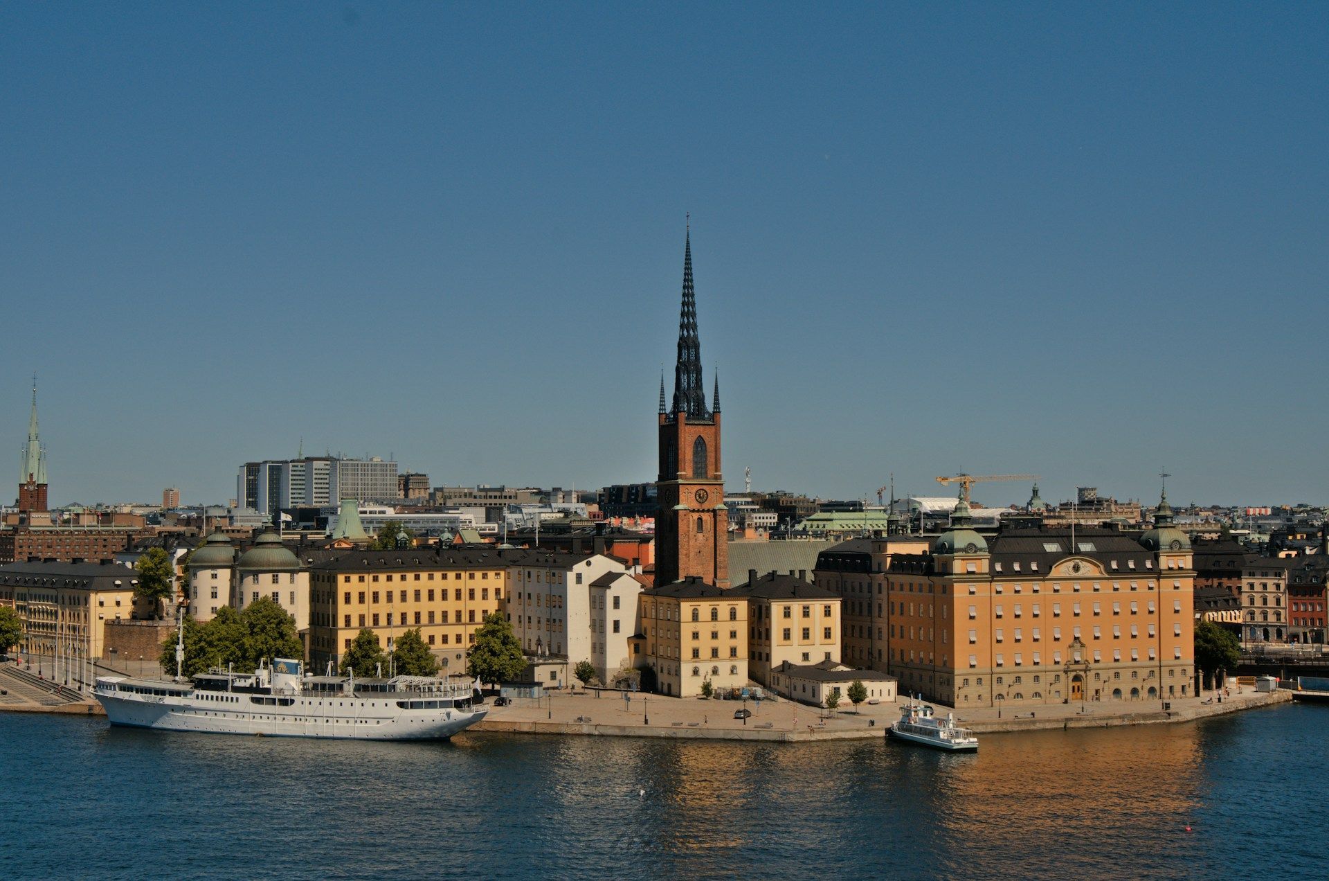 The Riddarholmen Church spire rises above historic buildings on the waterfront in Stockholm, Sweden, under a clear sky.