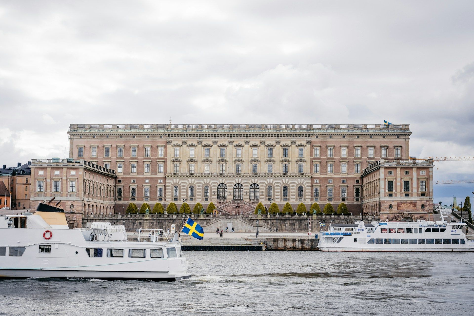 The Stockholm Palace stands on the waterfront with boats in the foreground under a cloudy sky.