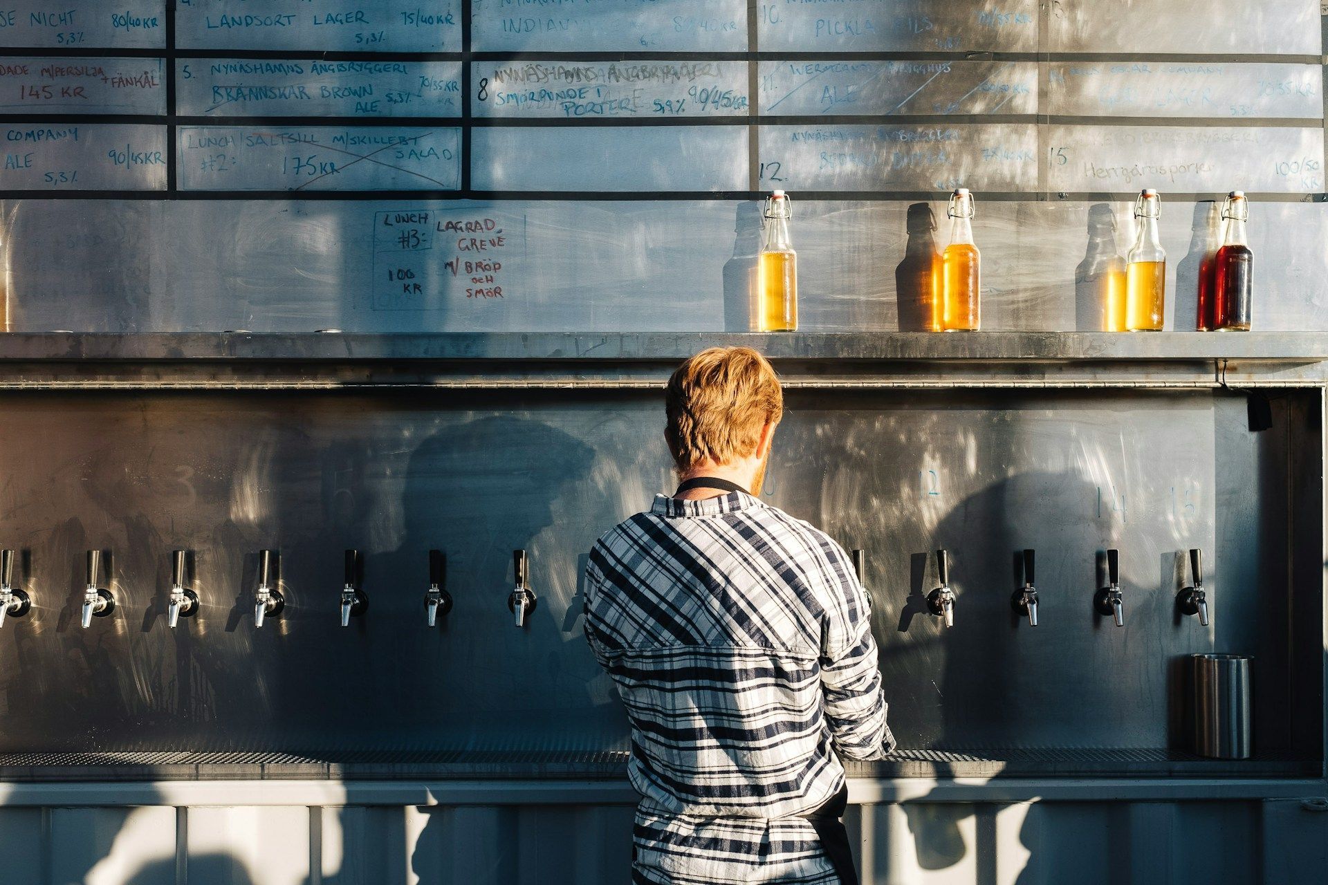 A person in a plaid shirt stands before a metal wall of beer taps in Stockholm.
