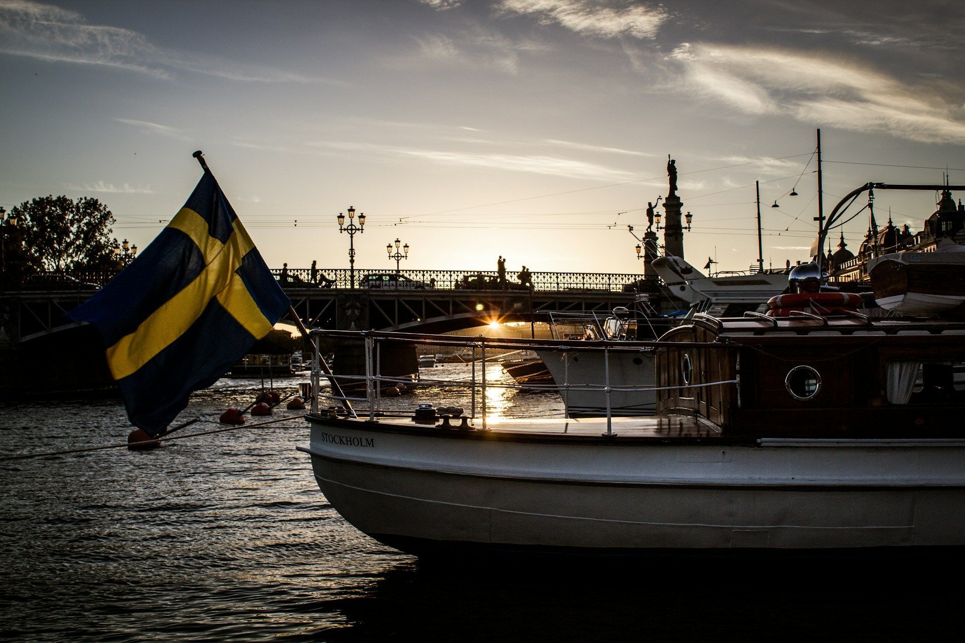 A Swedish flag flies from the stern of a boat on a calm river at sunset.