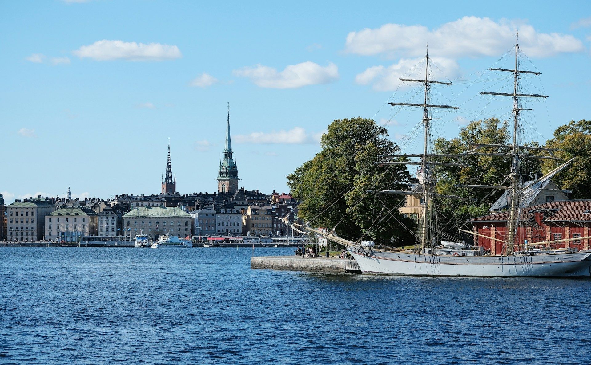 A tall-masted ship docked at a pier with the historic skyline of Gamla Stan in Stockholm in the background.