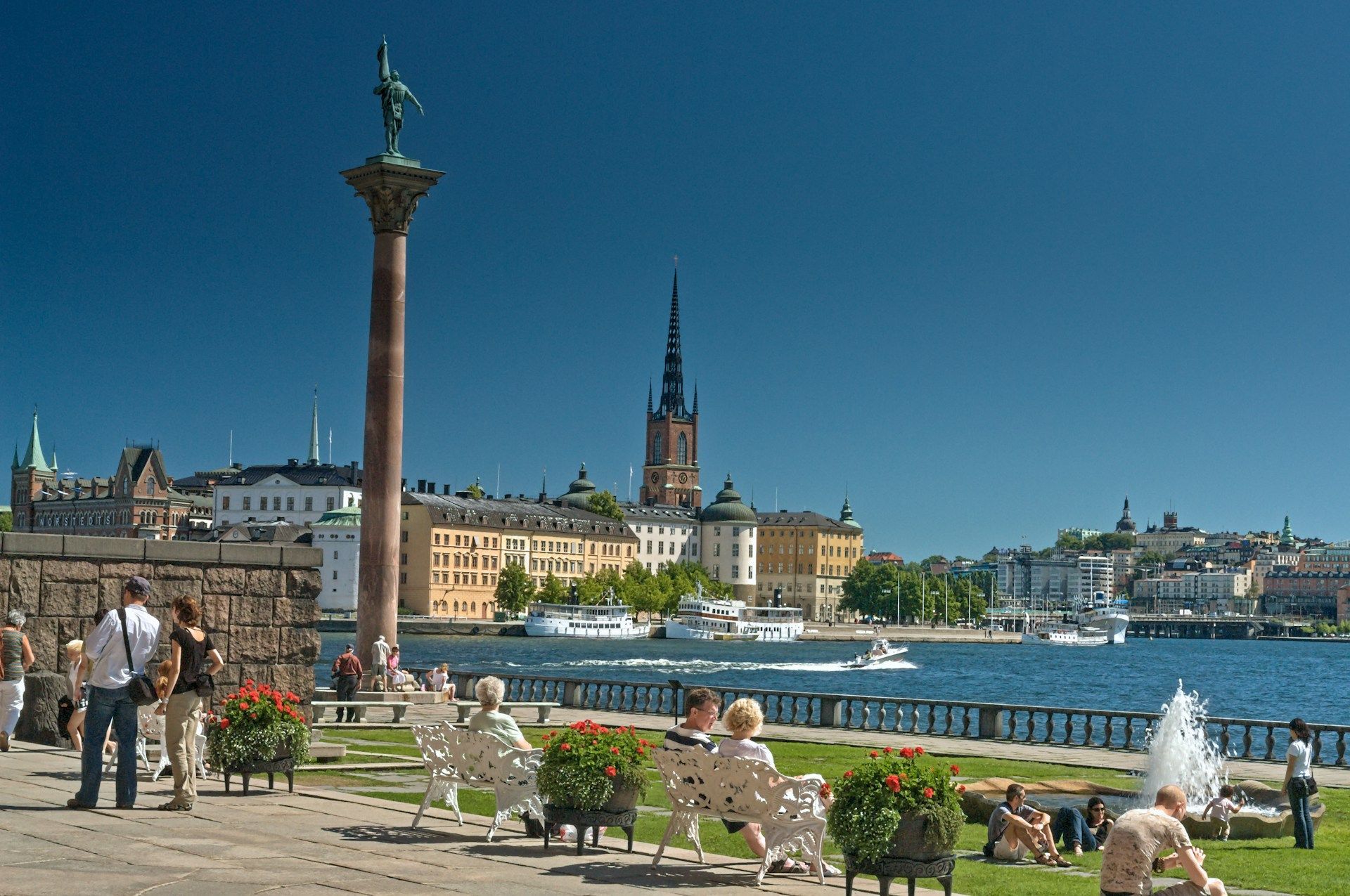 People relax at Stockholm City Hall along the waterfront, featuring Riddarholmen Church in the background.