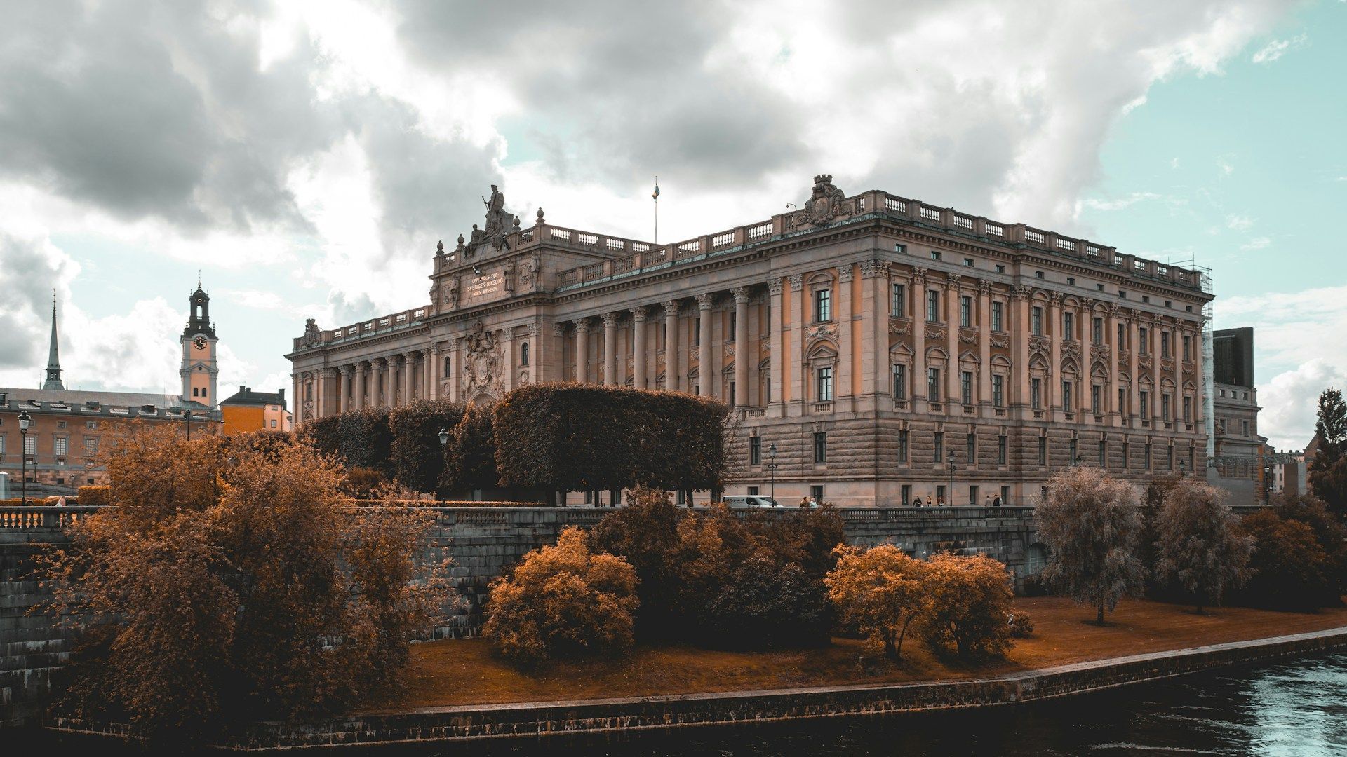 The Swedish Parliament building (Riksdagshuset) in Stockholm, viewed from across the water with autumn trees in the front.