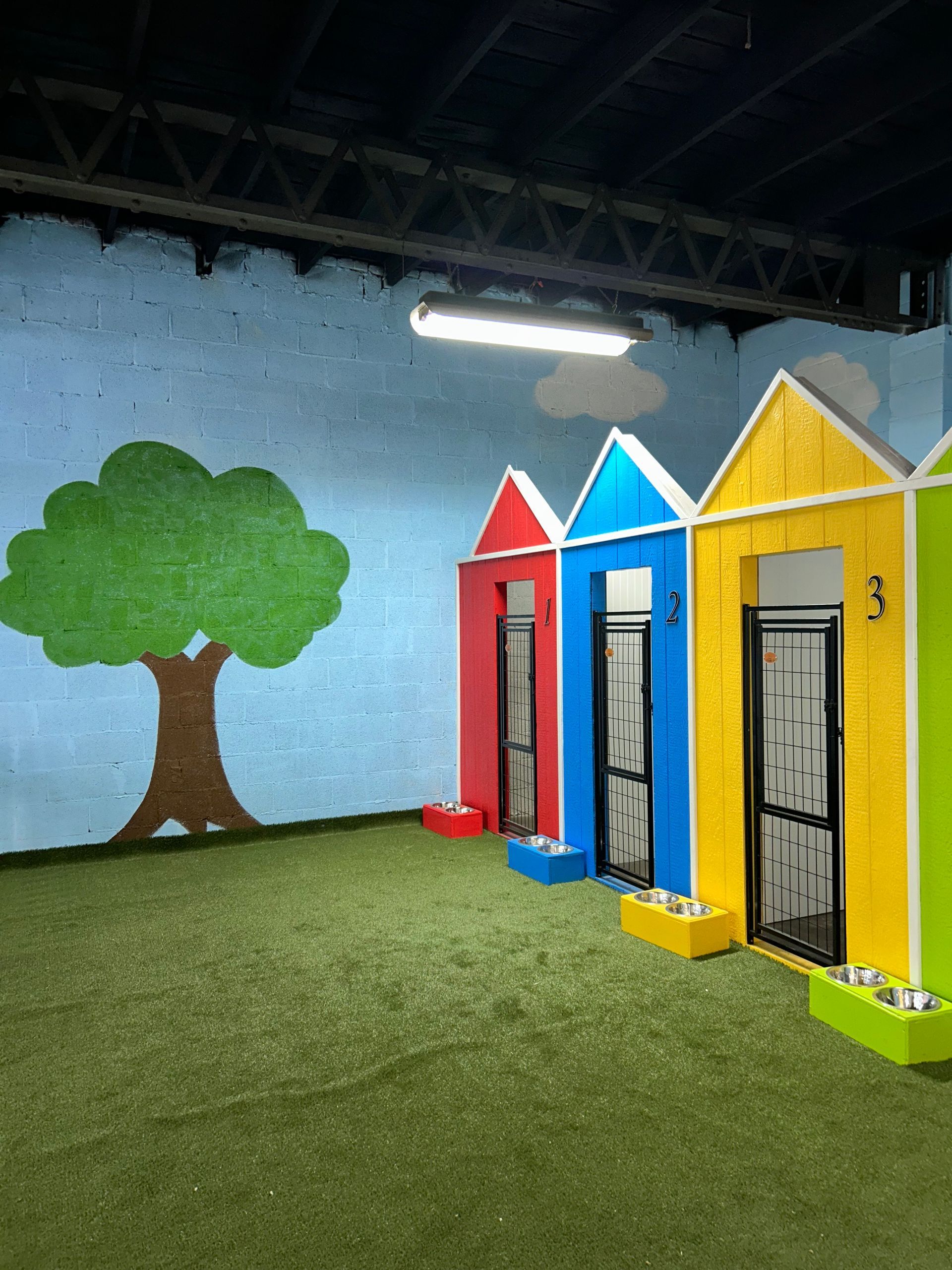 Colorful dog kennels with house-shaped roofs, a painted tree, and artificial grass.