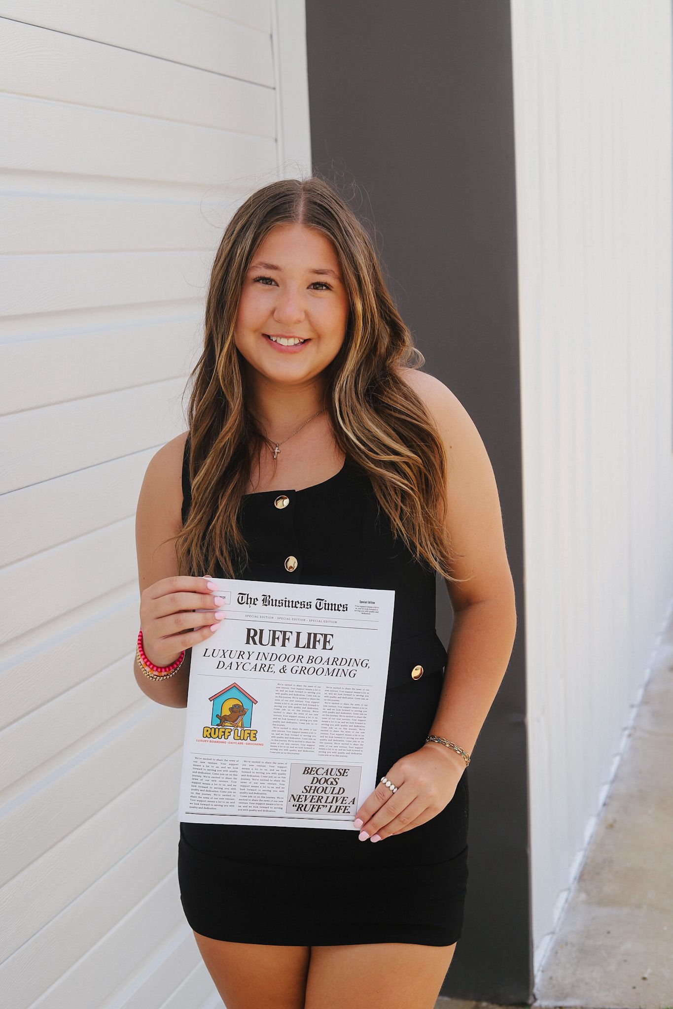 Woman in black dress holds a certificate, smiling outside against white and gray walls.