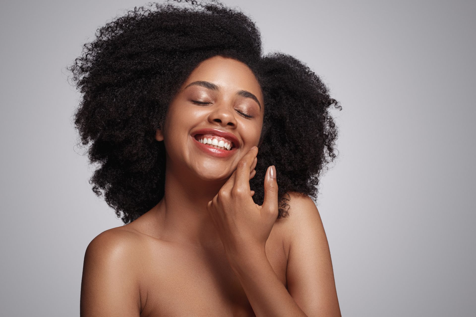 A woman with curly hair is smiling and touching her face.