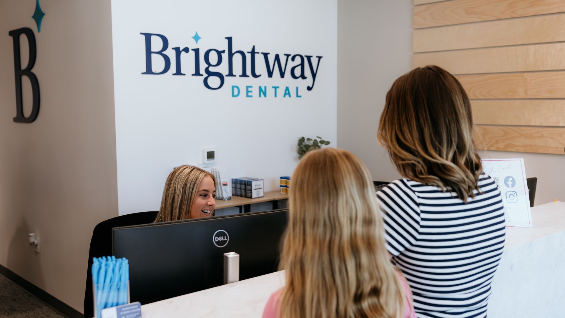 At a dental office, a receptionist smiles at a woman and child standing at the front desk, beneath a