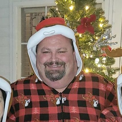 A man wearing a santa hat and a plaid shirt is standing in front of a christmas tree.