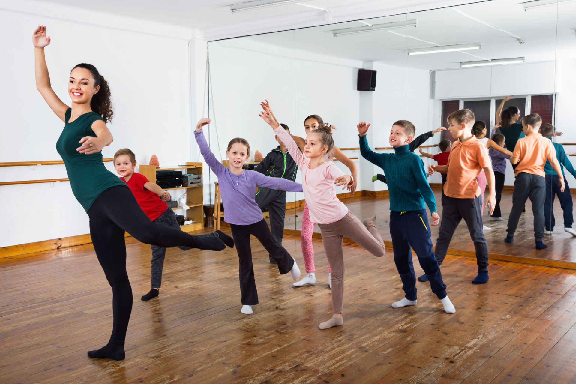 People dancing in a bright studio, posing with raised arms and legs on a wooden floor.