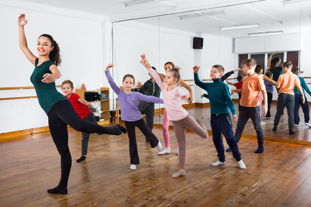 People dancing in a bright studio, posing with raised arms and legs on a wooden floor.
