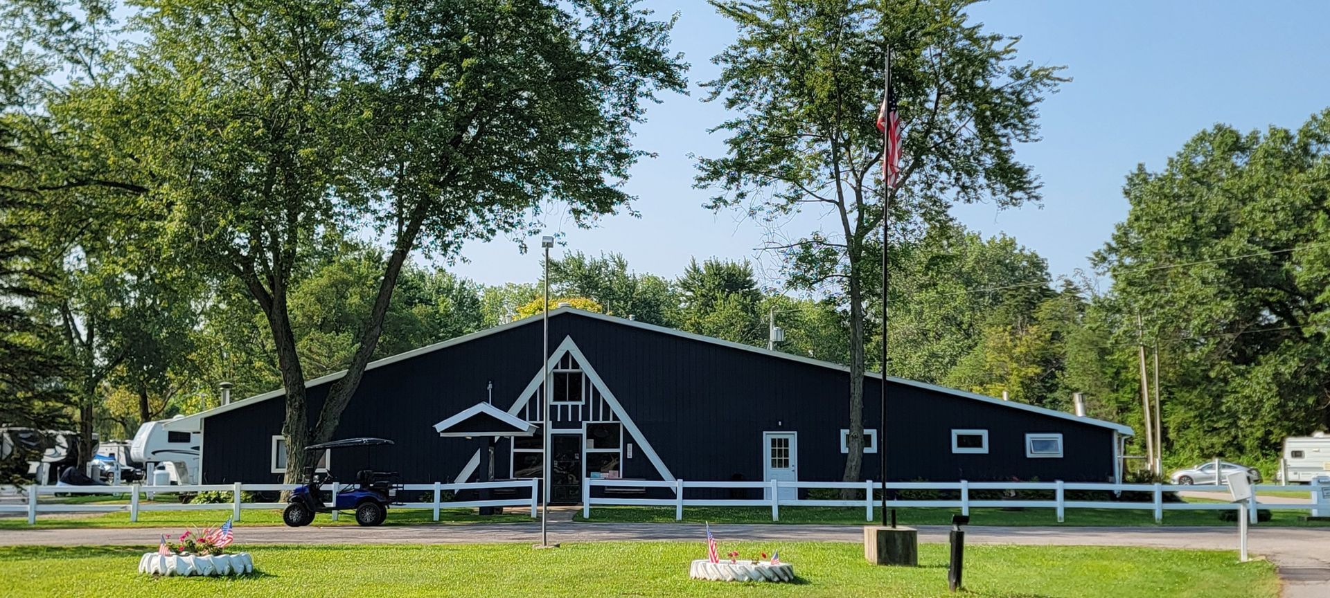A large black barn with a white fence and trees in front of it.