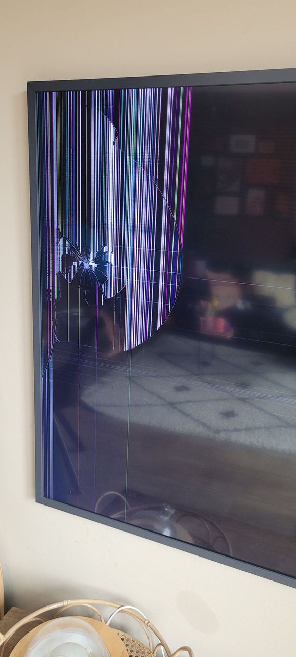 Een grote televisie aan de muur met een gebroken scherm, waarop kleurrijke strepen en vervorming te zien zijn. In de onderste hoek van het scherm zijn wat glasscherven te zien.