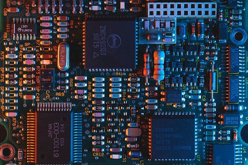 Close-up van een groene printplaat vol met diverse elektronische componenten. Oranje en blauwe verlichting accentueert de complexe details.