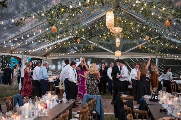 Wedding reception under a clear tent with string lights and dancing guests. Tables are set with floral centerpieces.