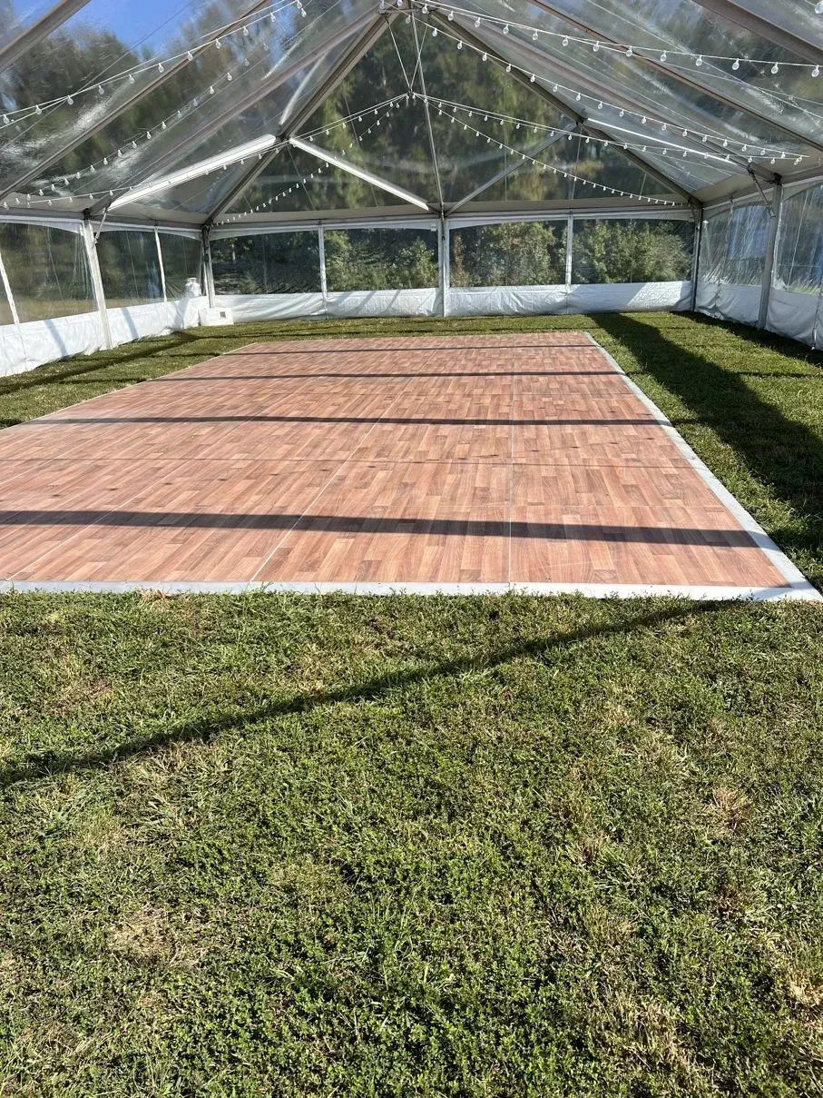 A dance floor, wood-look, is set on grass inside a clear tent. White edging.