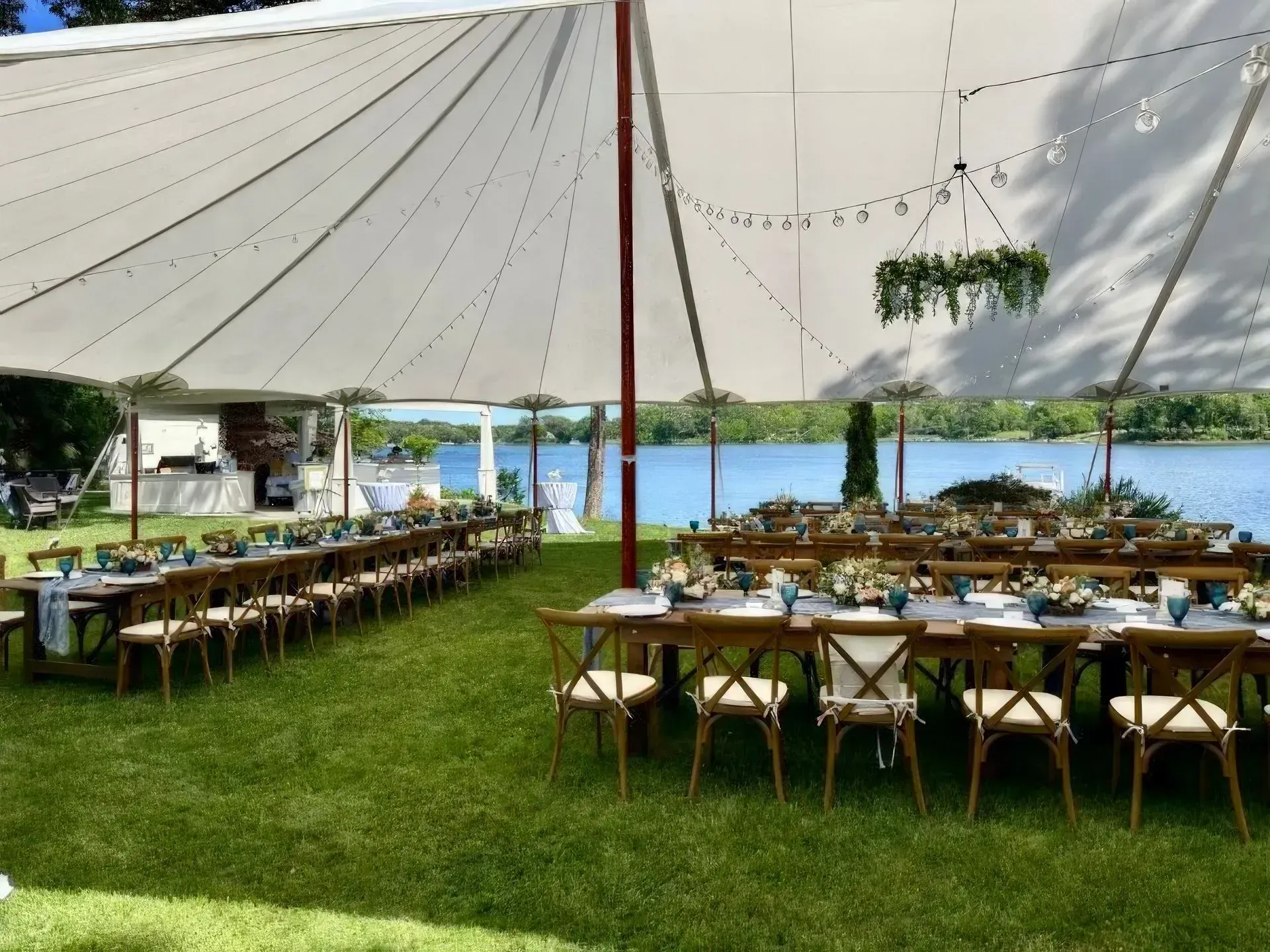 Wedding reception setup under a white tent on a grassy lawn overlooking a lake; tables set for guests.
