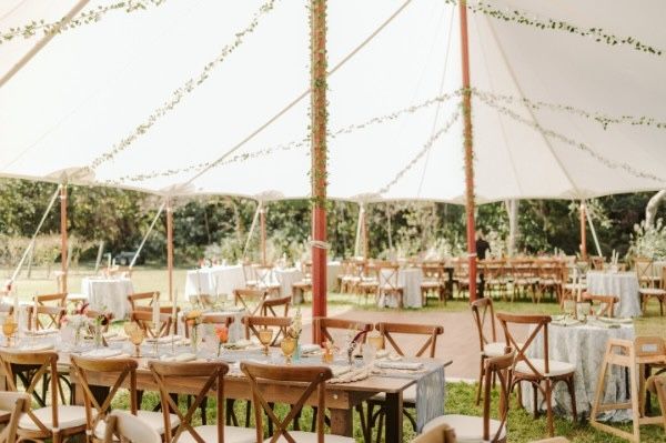 Wedding reception tent with wooden tables, chairs, and white tablecloths, set outdoors.
