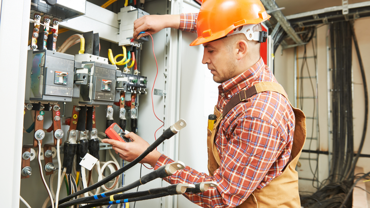 Electrician in orange hard hat working on electrical panel, using a multimeter.