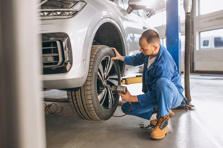 Mechanic in blue coveralls, crouching by a car wheel, using a wrench in a garage.