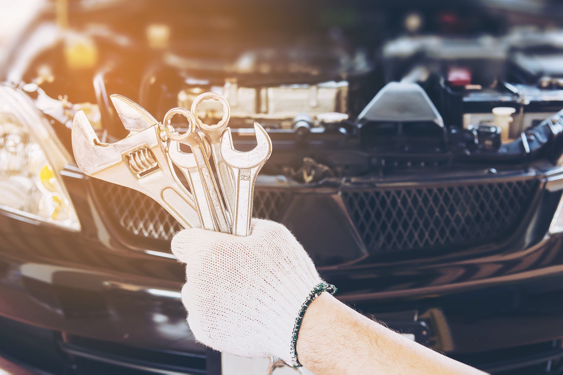 Gloved hand holding wrenches in front of an open car hood.