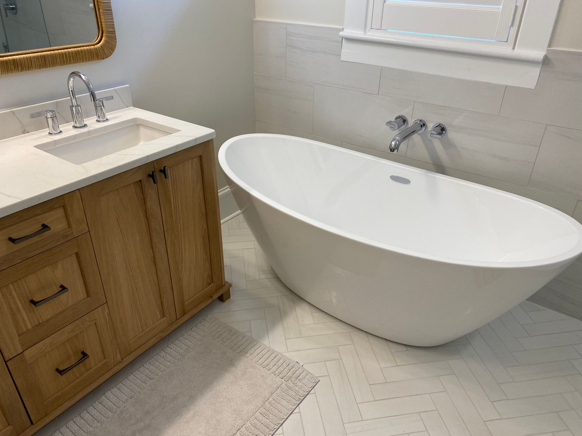 Custom white oak vanity cabinets next to a freestanding oval tub with a wall mount filler and herringbone laid tile floor.