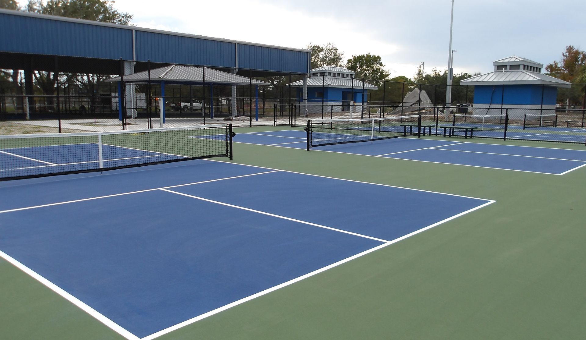 Blue tennis court with white lines and net, surrounded by a green area and blue fencing, trees in the background.
