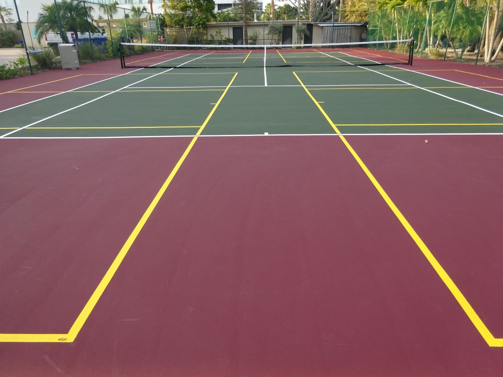 A chain-link fence separates a sports court with a red and green surface from a wooded area with fall foliage.