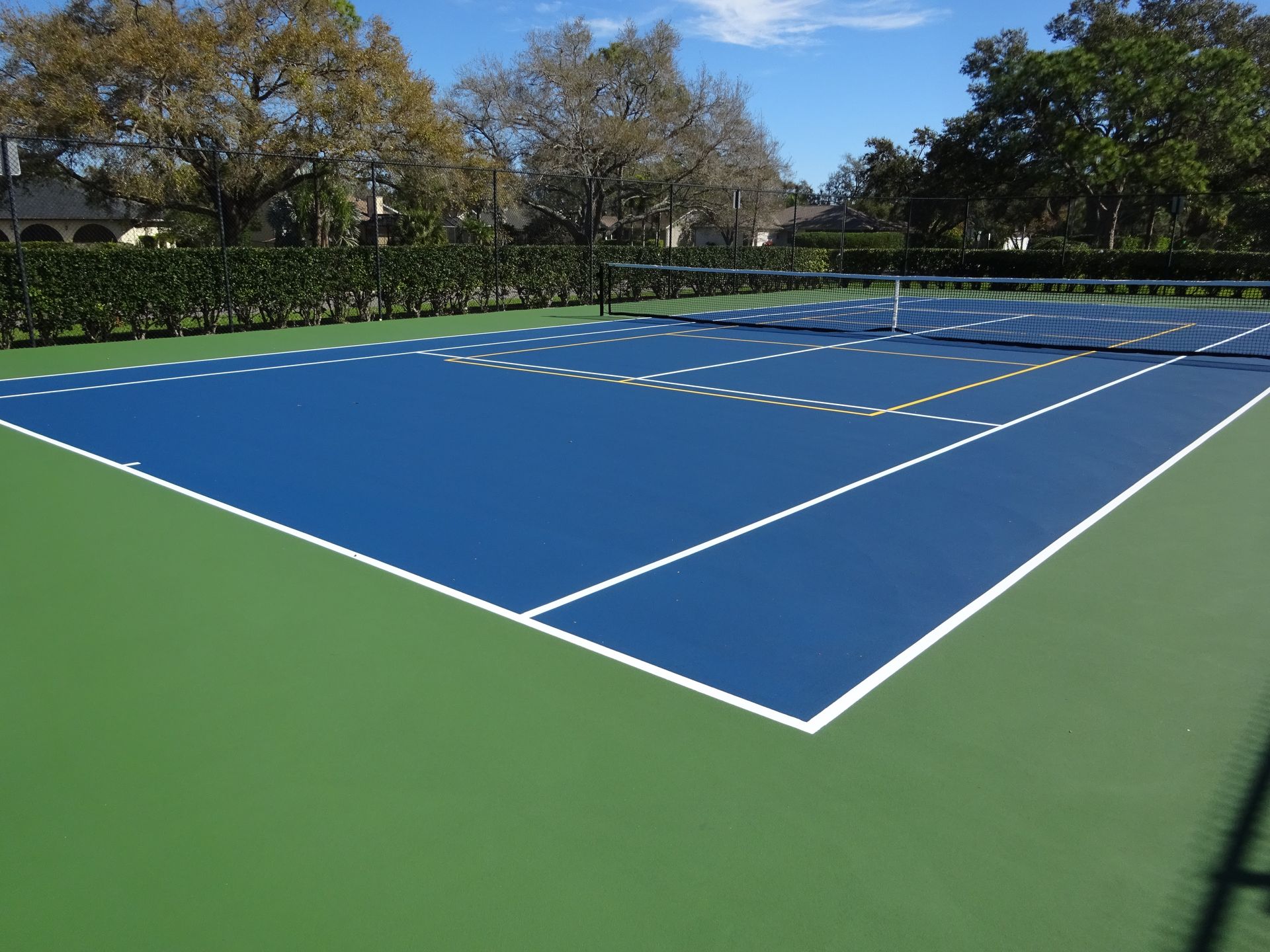 Blue tennis court with white lines and net, surrounded by a green area and blue fencing, trees in the background.
