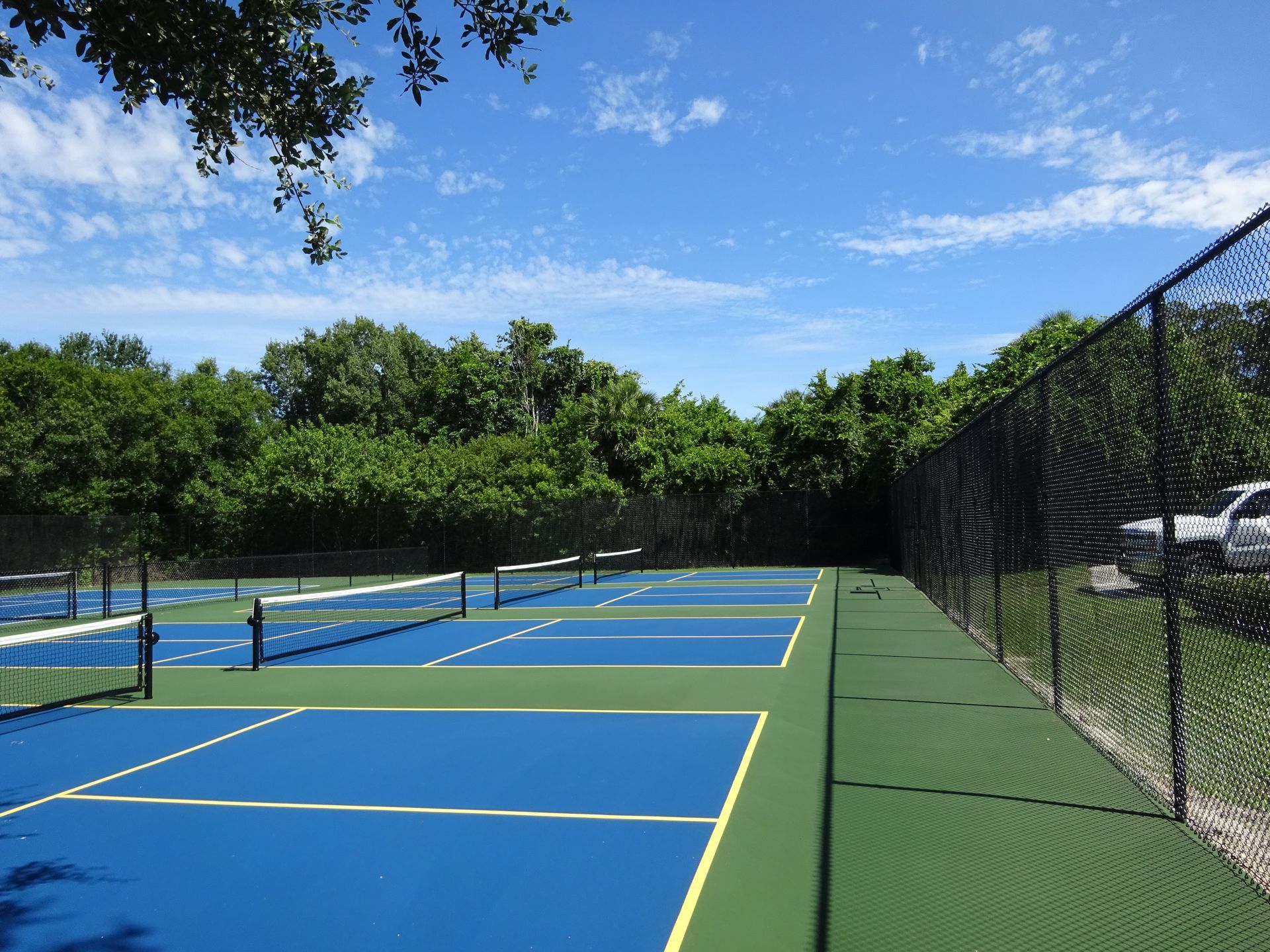 Blue tennis court with white lines and net, surrounded by a green area and blue fencing, trees in the background.