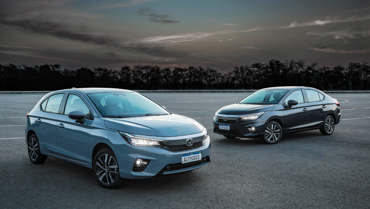 Dois carros Honda City, um hatchback e um sedan, estacionados no asfalto. Carro azul na frente, carro cinza escuro ao fundo. Céu nublado.