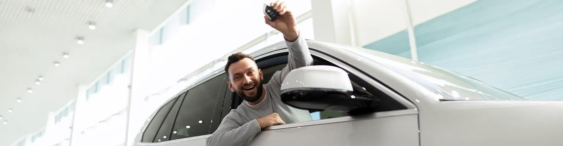 Homem sorrindo da janela de um carro prateado, levantando o braço dentro de um showroom iluminado.