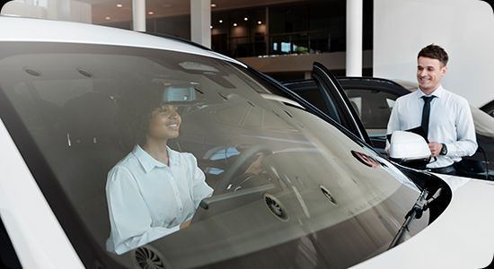 Um vendedor de carros está ao lado de um carro branco enquanto uma pessoa está sentada dentro, vista através do para-brisa.