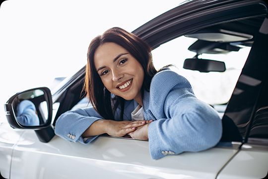 Mulher sorrindo e debruçada para fora da janela de um carro branco, vestindo uma camisa azul e com os braços apoiados na porta.