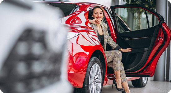 Mulher sentada na porta aberta de um carro vermelho, sorrindo e falando ao telefone em uma concessionária.