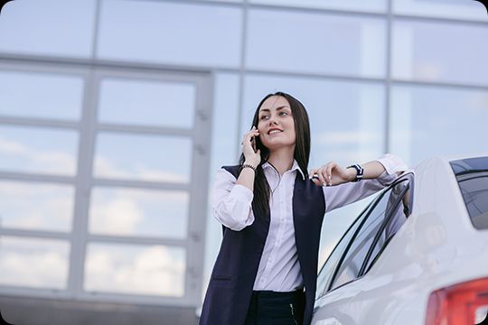 Mulher ao telefone ao lado de um carro branco em frente a um prédio moderno, sorrindo e encostada no veículo.