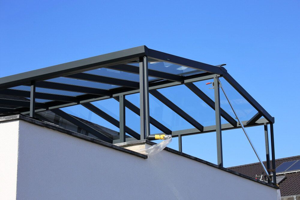 Glass-Roofed Patio with Dark Gray Frame and White Wall Against a Clear Blue Sky — Mingweld Fabrication in Bungalow, QLD