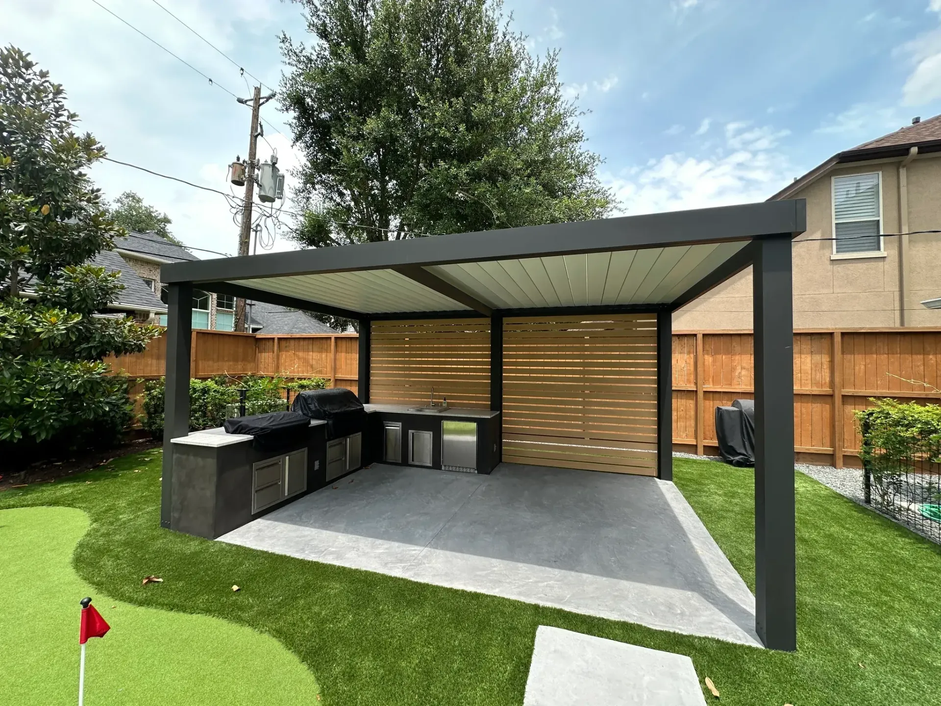 Outdoor kitchen under pergola with grill, sink, and built-in cabinets; green grass and backyard setting.