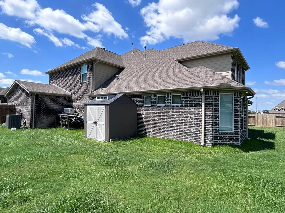 Backyard of a brick house with a shed on a grassy lawn under a blue sky with clouds.