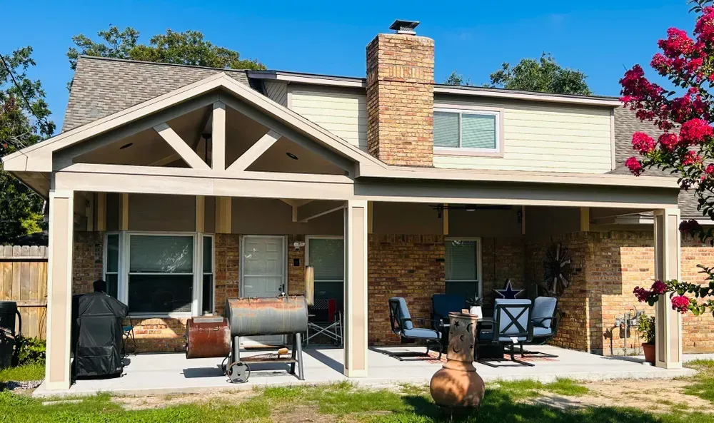 Backyard patio with brick, seating, and a chimney, under a two-story house with blue sky.