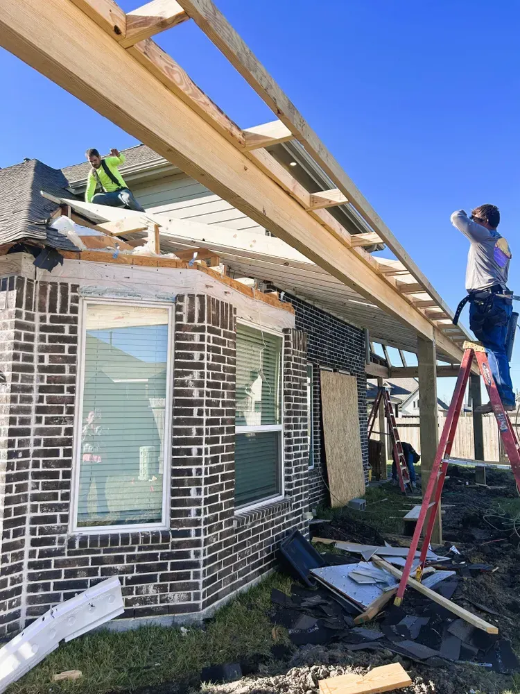 Construction workers building a covered patio attached to a brick house. Bright blue sky overhead.