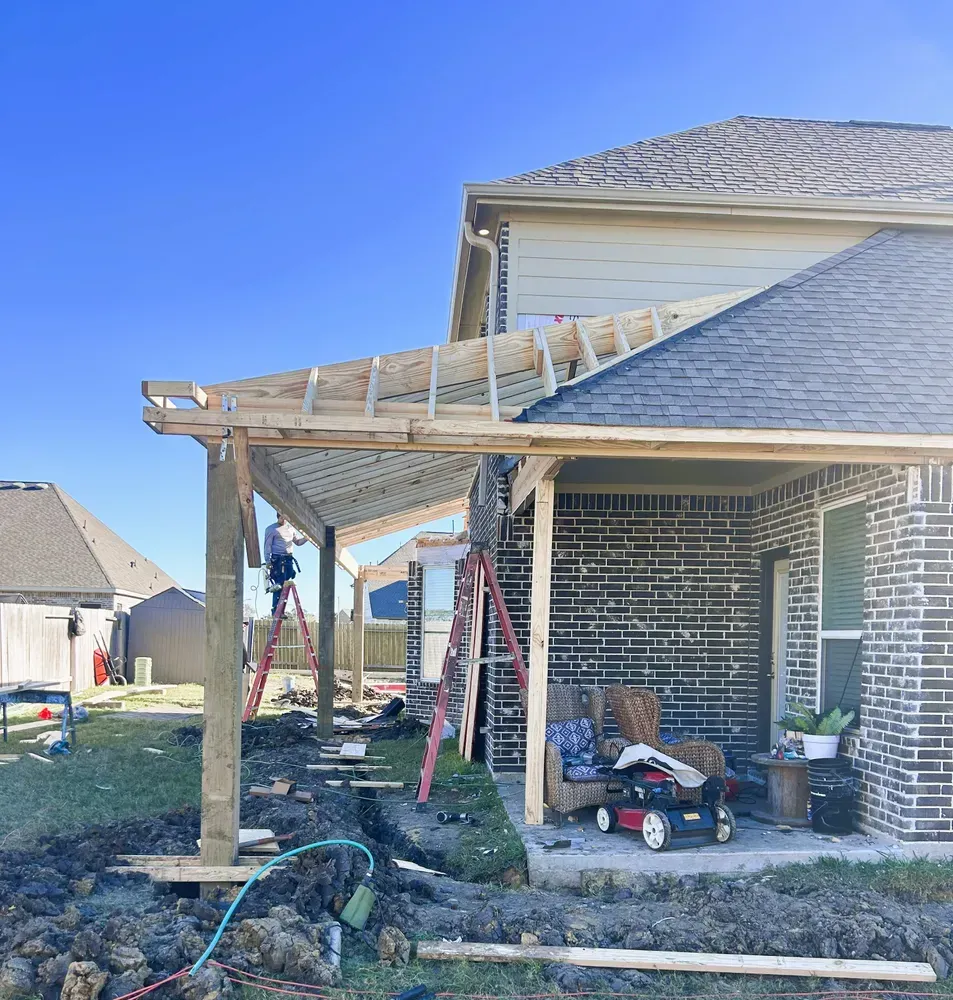 Construction of a wooden patio cover attached to a brick house; workers on ladders.