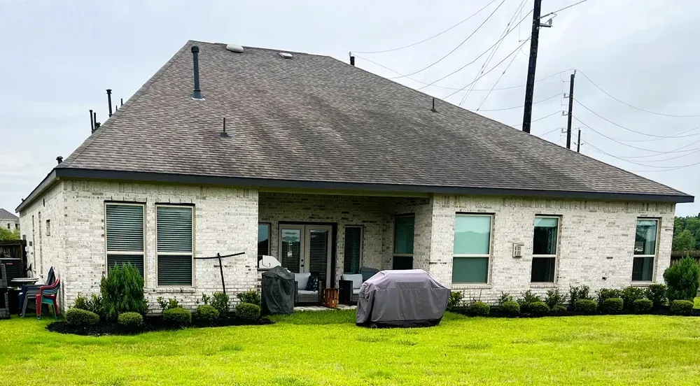 Back of a brick house with dark roof, glass doors, windows, green grass, and bushes.