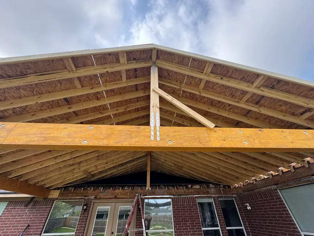 Construction of a covered patio with exposed wood beams, supported by a brick home, under a cloudy sky.