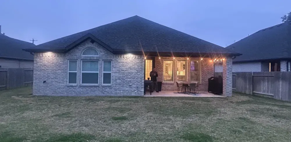 Backyard view of a brick house with a lit patio, person standing, and string lights. Overcast sky.