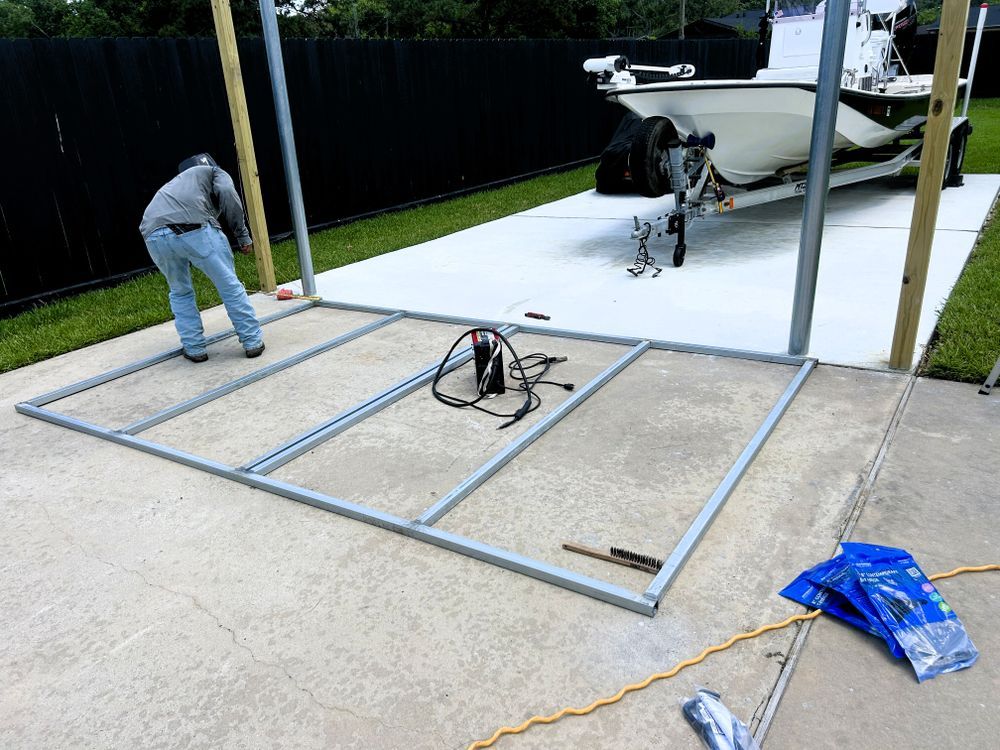 Man assembling metal carport frame on concrete driveway with boat in background.
