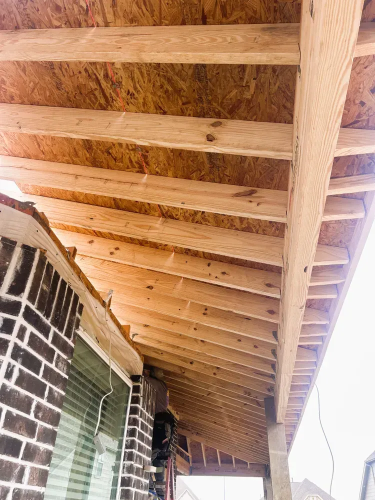Wooden roof structure under construction, adjacent to a brick wall with a window.