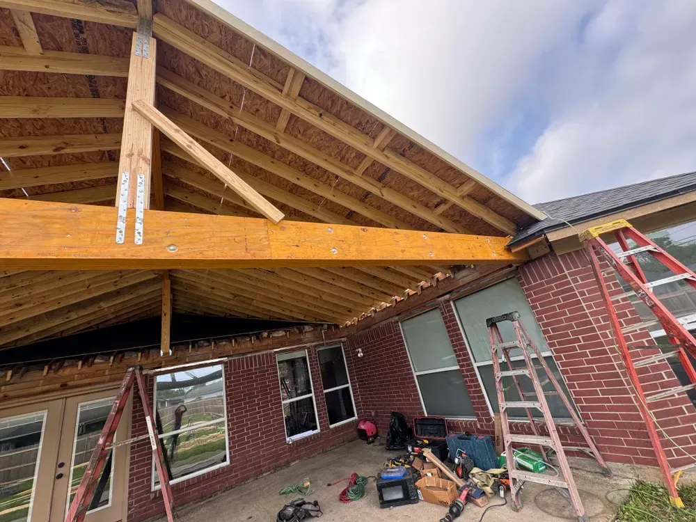 A wooden patio roof being built over a brick house, with tools and ladders on the ground.