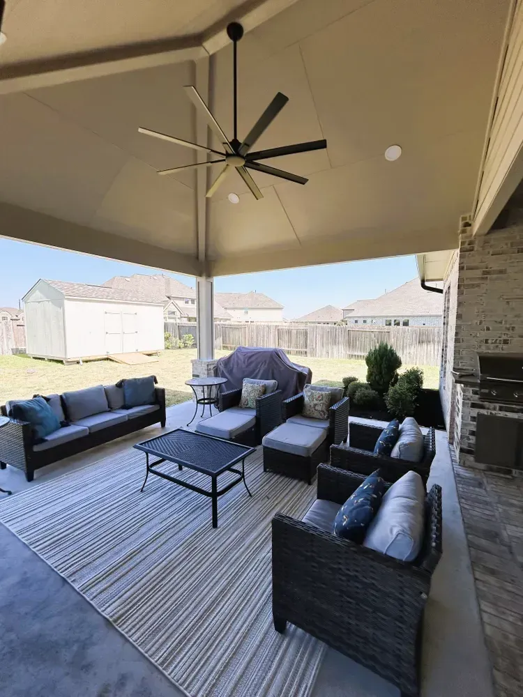 Covered patio with outdoor furniture: gray sofa, wicker chairs, rug, and ceiling fan. View of yard.