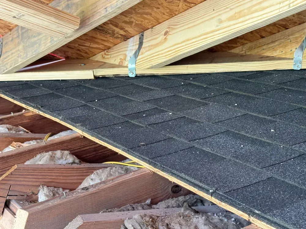 Partially installed shingle roof, showing black shingles, wooden trusses, and insulation.