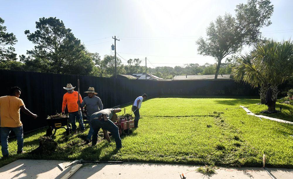 Men working in a grassy backyard, using tools to landscape. Bright sunlight. Fence and trees in background.